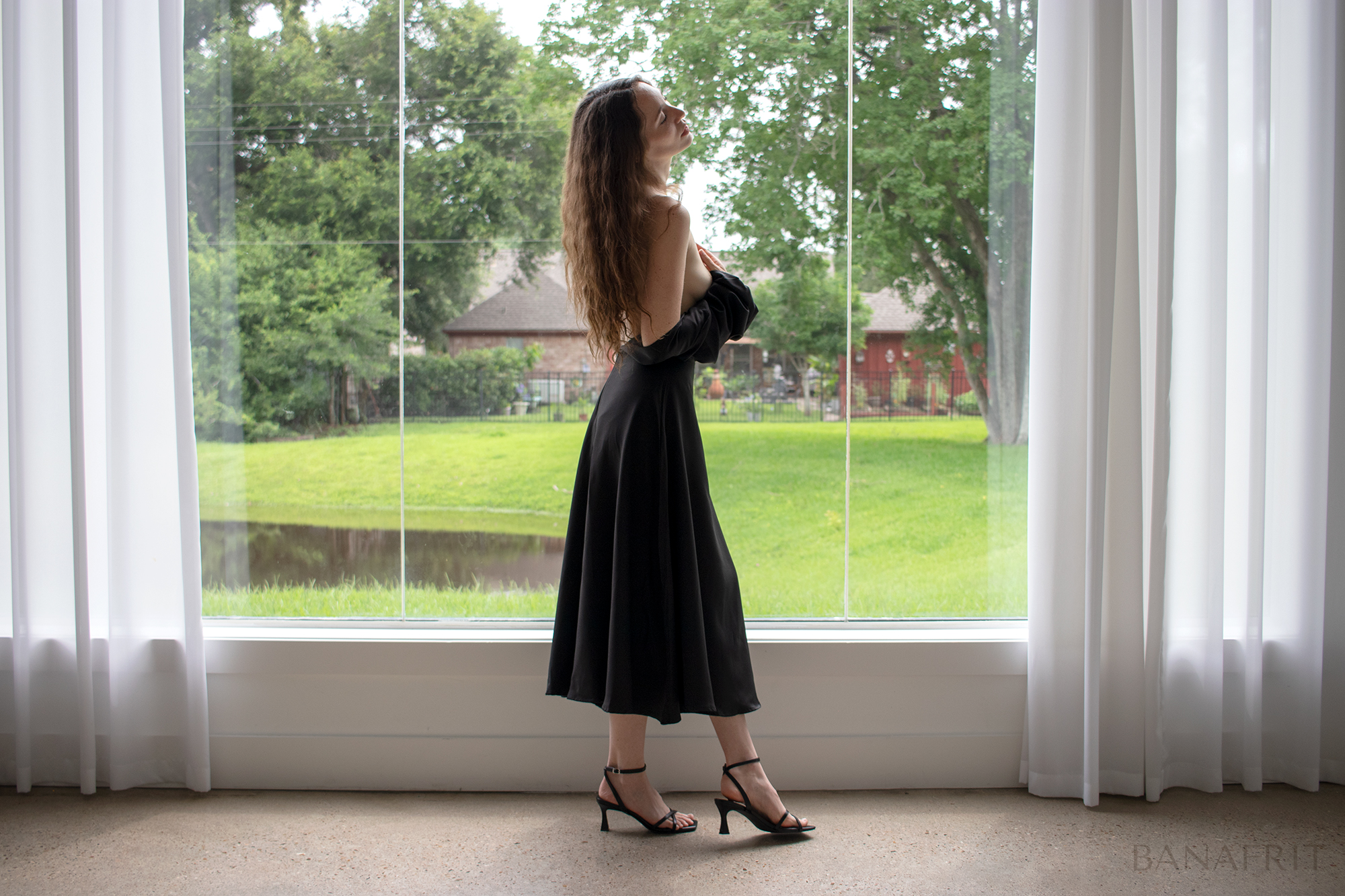 Woman in backless black dress and heels standing in profile by a large window overlooking a garden
