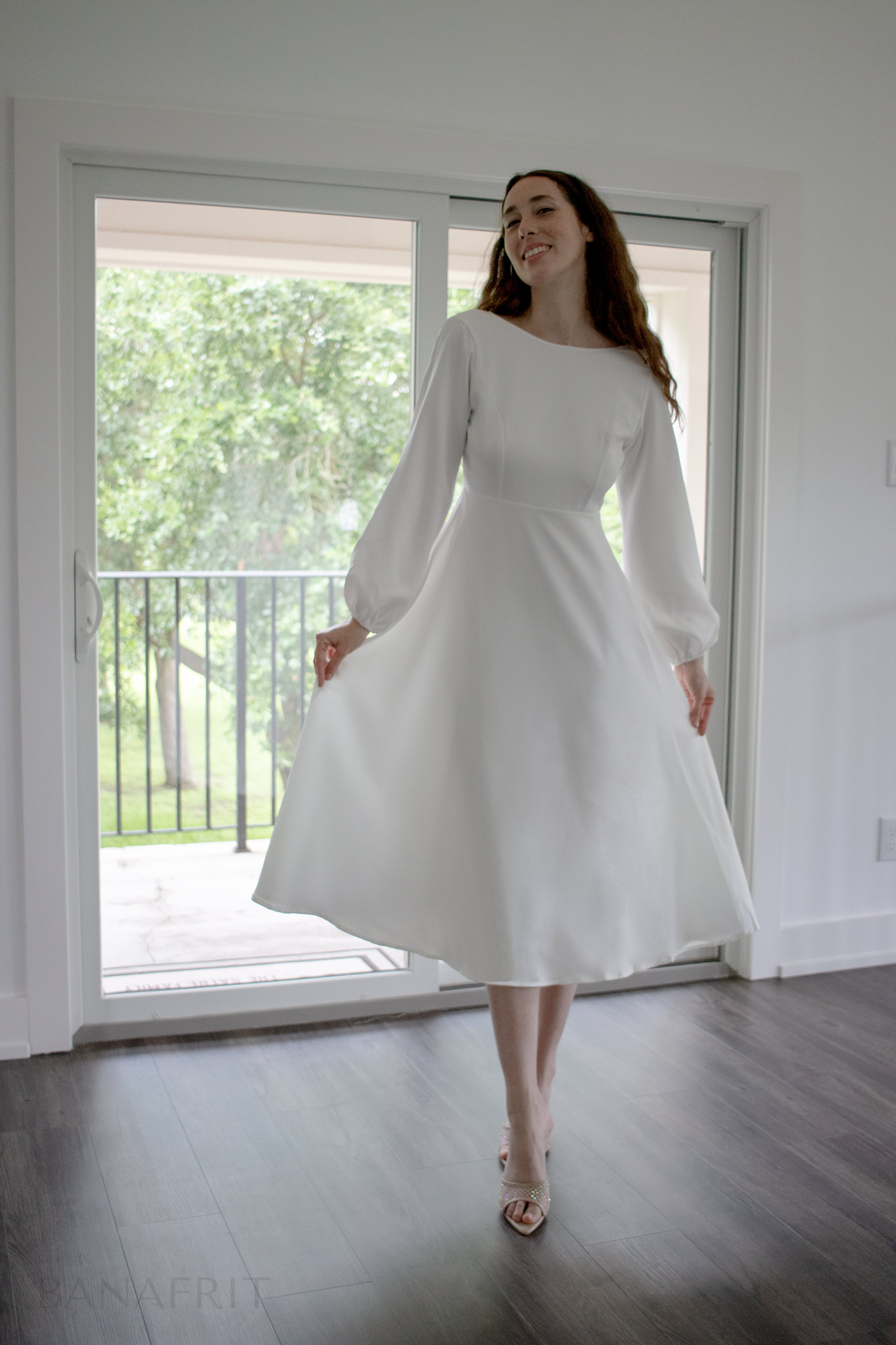 Smiling woman in elegant long-sleeve white midi dress twirling by glass doors